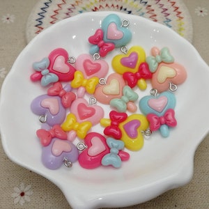 May include: A collection of 14 colorful heart-shaped charms with bows. Each charm has a silver loop for attaching to jewelry or other crafts.
