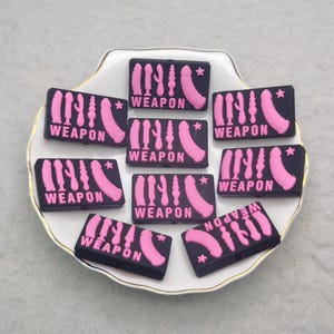 May include: A collection of black rectangular patches with pink designs and the word "WEAPON". The patches feature various stylized phallic shapes and a curved design, arranged on a white plate.