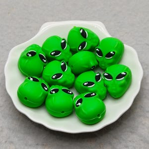 Puede incluir: Un conjunto de cuentas verdes con forma de cabeza de alienígena con ojos negros y blancos, dispuestas sobre un plato blanco con borde festoneado. Las cuentas son de color verde vibrante, y los ojos tienen un diseño detallado.