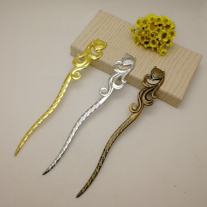 Puede incluir: Tres palitos decorativos para el pelo con diseños ornamentados. Los palitos son de color dorado, plateado y bronce y presentan una cabeza de animal estilizada en la parte superior.