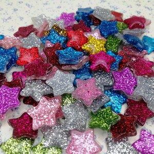 Puede incluir: Un gran surtido de adornos con forma de estrella de purpurina en varios colores, incluyendo plata, oro, rojo, azul, rosa y verde.