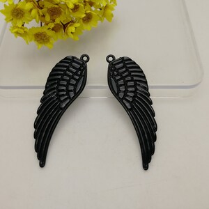 以下が含まれることがあります： 光沢のある黒い金属製の翼チャーム2つ。各翼には詳細な羽毛があり、ジュエリーやその他の工芸品に取り付けるための小さなループが上部にあります。翼は白い表面に表示されています。