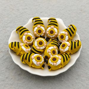 May include: A collection of yellow and black gnome figurines with sunflower details. The gnomes are arranged on a white, scalloped-edge plate. The figurines have striped hats and bodies, and are approximately 1 inch in height.