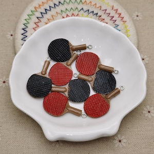 May include: A group of eight miniature ping pong paddle charms in red and black with brown handles. The charms are on a white plate.