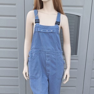 Op de afbeelding: Een blauwe denim overall met zwarte banden en een knoopsluiting. De overall heeft twee grote zakken aan de voorkant en een kleinere zak op de borst.