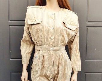 May include: A beige striped jumpsuit with long sleeves and a button-up front. The jumpsuit has a fitted bodice and wide-leg pants.