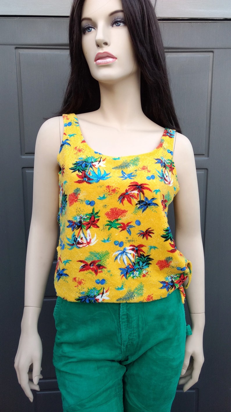 May include: A yellow tank top with a tropical print of palm trees, flowers, and leaves. The tank top has a scoop neck and thin straps. The tank top is made of a soft, textured fabric.