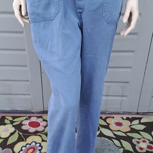 Op de afbeelding: Een blauwe denim overall met een knoopsluiting en twee voorzakken. De overall is op een mannequin en staat op een bloemenkleed.
