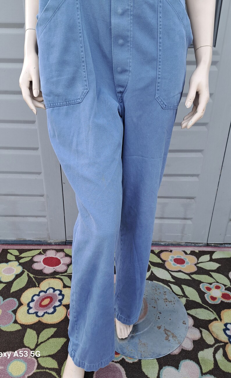 Op de afbeelding: Een blauwe denim overall met een knoopsluiting en twee voorzakken. De overall is op een mannequin en staat op een bloemenkleed.