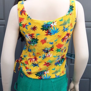 May include: A yellow tank top with a tropical print. The tank top has thin straps and a scoop neck. The tank top is paired with green corduroy pants.