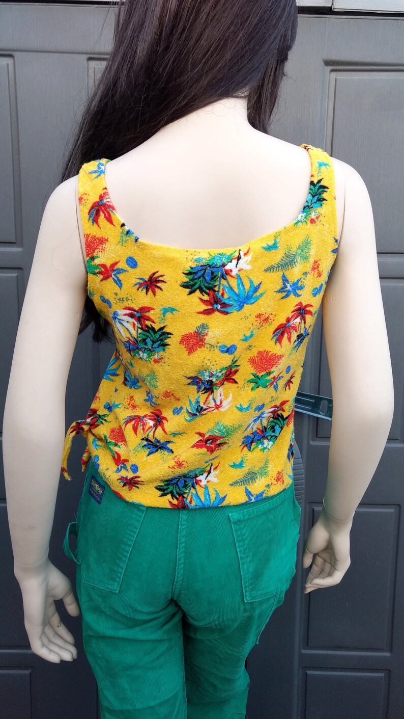 May include: A yellow tank top with a tropical print. The tank top has thin straps and a scoop neck. The tank top is paired with green corduroy pants.