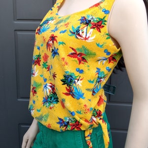 May include: A yellow sleeveless top with a tropical print featuring palm trees, flowers, and leaves. The top has a tie at the waist and is made of a soft, textured fabric.