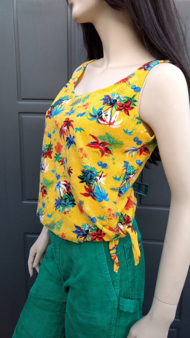 May include: A yellow sleeveless top with a tropical print featuring palm trees, flowers, and leaves. The top has a tie at the waist and is made of a soft, textured fabric.
