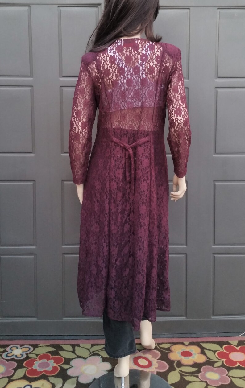Puede incluir: Una larga chaqueta de encaje color burdeos con un lazo en la espalda. La chaqueta es transparente y tiene un patr&oacute;n floral.