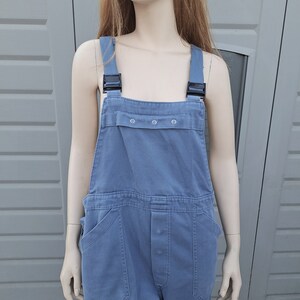 Op de afbeelding: Een blauwe denim overall met zwarte banden en een knoopsluiting. De overall heeft twee grote zakken aan de voorkant en een klein zakje op de borst.