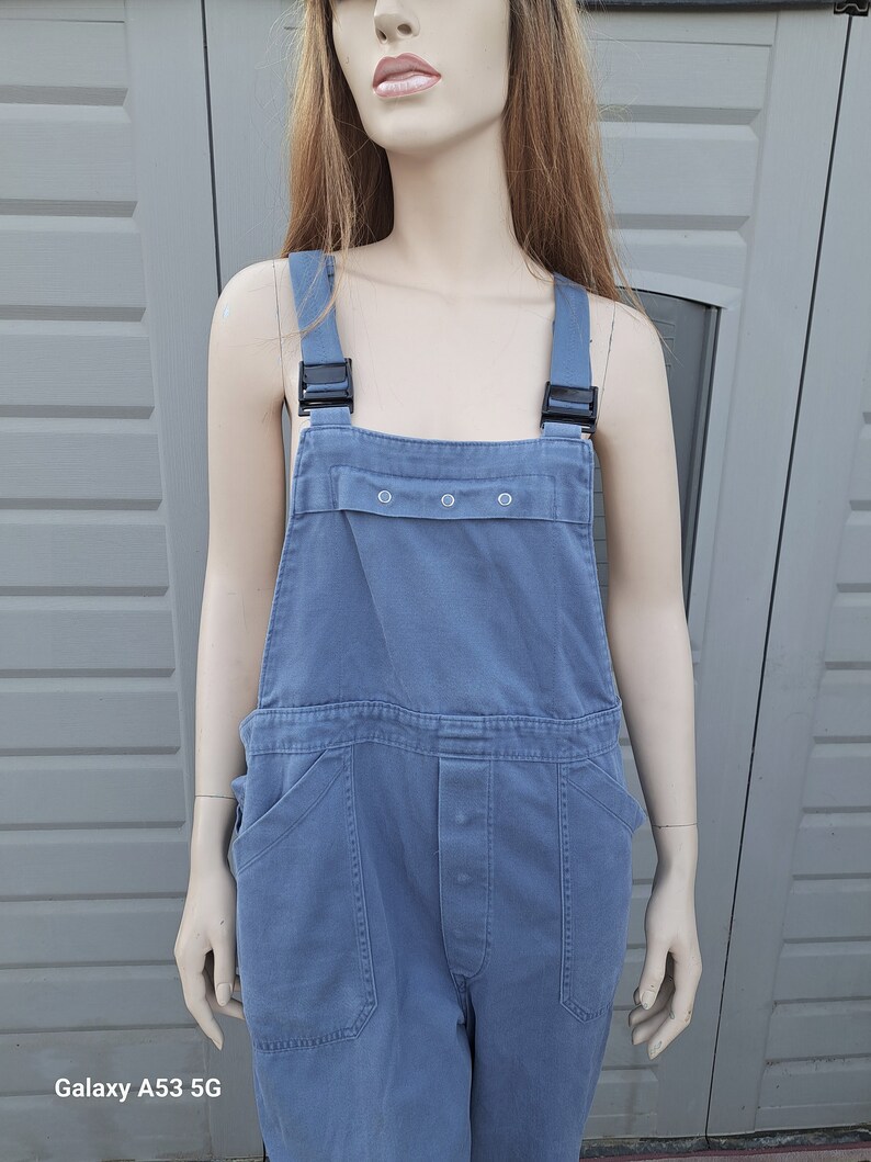 Op de afbeelding: Een blauwe denim overall met zwarte banden en een knoopsluiting. De overall heeft twee grote zakken aan de voorkant en een klein zakje op de borst.