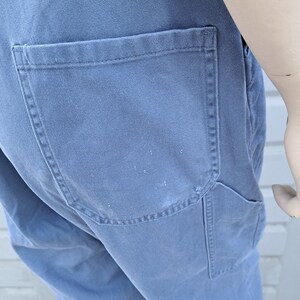 Op de afbeelding: Een blauwe denim overall met een grote achterzak. De overall heeft een verweerde look en is gemaakt van een stevige stof.