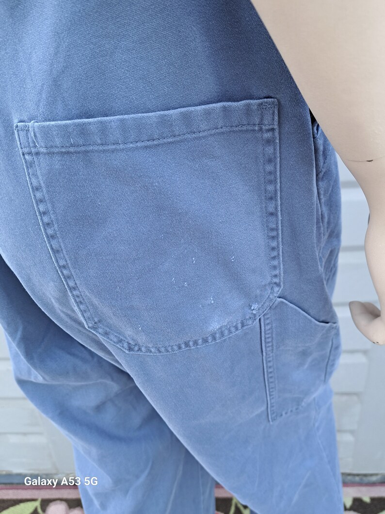Op de afbeelding: Een blauwe denim overall met een grote achterzak. De overall heeft een verweerde look en is gemaakt van een stevige stof.