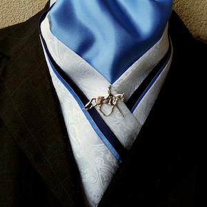 May include: A white and blue patterned cravat with a blue satin top layer and a navy blue trim. A silver horse rider pin is attached to the cravat. The cravat is worn over a black suit jacket. The text "Doc's Designs" is at the bottom of the image.