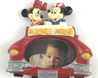 May include: A red Mickey Mouse car-shaped picture frame featuring Mickey and Minnie Mouse figurines. The frame holds a photograph of a baby. The words "Mickey Mouse" and "Mick N Min" are visible on the frame.