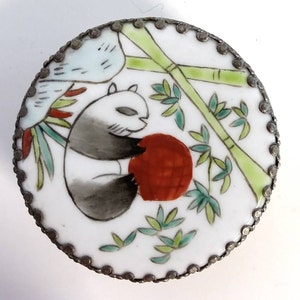 May include: A round porcelain trinket box with a silver metal rim. The lid features a hand-painted design of a panda bear holding a red ball, surrounded by bamboo stalks and green leaves.