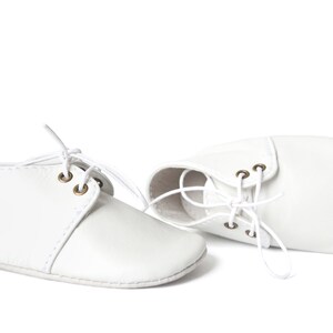 Op de afbeelding: Een paar witte babyschoentjes met veters. De schoenen zijn gemaakt van een zacht materiaal en hebben een klassiek ontwerp met oogjes voor de veters. De schoentjes zijn ontworpen voor baby's.