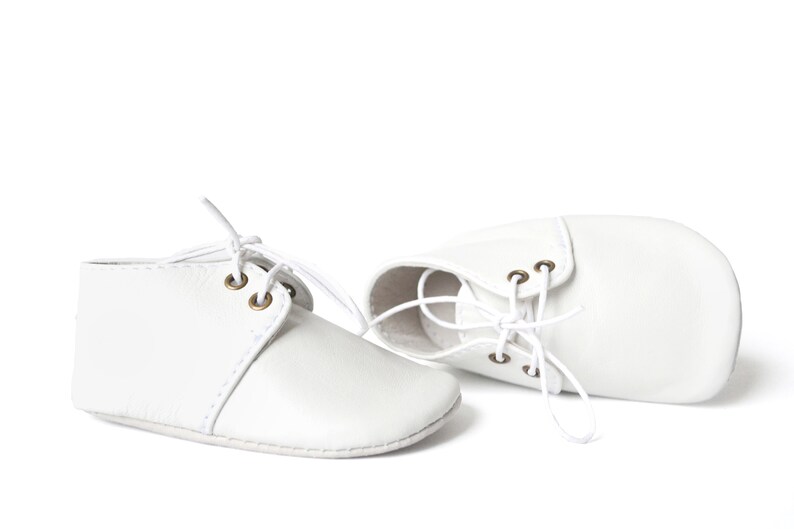 Op de afbeelding: Een paar witte babyschoentjes met veters. De schoenen zijn gemaakt van een zacht materiaal en hebben een klassiek ontwerp met oogjes voor de veters. De schoentjes zijn ontworpen voor baby's.