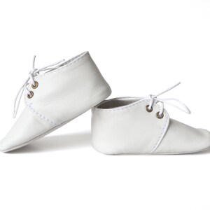 Op de afbeelding: Een paar witte leren babyschoenen met veters. De schoenen hebben een klassiek ontwerp met een ronde neus en een gestikt naaddetail. De vetergaten zijn messingkleurig.