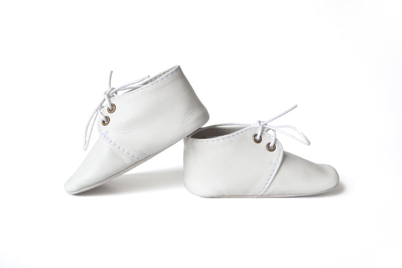 Op de afbeelding: Een paar witte leren babyschoenen met veters. De schoenen hebben een klassiek ontwerp met een ronde neus en een gestikt naaddetail. De vetergaten zijn messingkleurig.