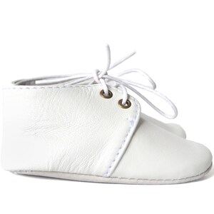 Op de afbeelding: Witte leren babyschoentjes met witte veters. De enkellaarsjes hebben een klassiek design met een gestikt naaddetail en messing oogjes. De veters zijn in een strik geknoopt.