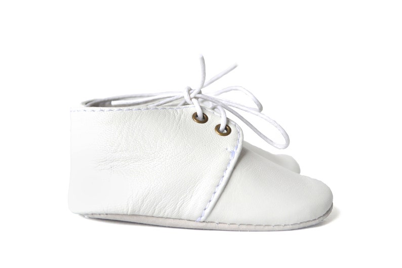Op de afbeelding: Witte leren babyschoentjes met witte veters. De enkellaarsjes hebben een klassiek design met een gestikt naaddetail en messing oogjes. De veters zijn in een strik geknoopt.