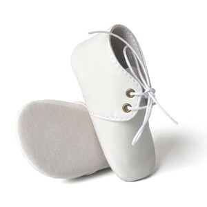 Op de afbeelding: Een paar witte babyschoenen. De schoenen zijn gemaakt van een glad materiaal en hebben een vetersluiting met witte veters en goudkleurige oogjes. De zolen zijn zacht off-white van kleur.