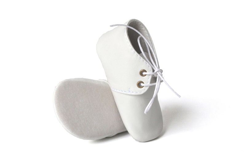 Op de afbeelding: Een paar witte babyschoenen. De schoenen zijn gemaakt van een glad materiaal en hebben een vetersluiting met witte veters en goudkleurige oogjes. De zolen zijn zacht off-white van kleur.