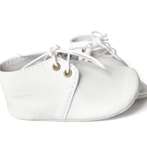 Op de afbeelding: Een paar witte leren babyschoenen met veters. De schoenen hebben een klassiek ontwerp met een ronde neus en kleine messing oogjes. De veters zijn wit en geknoopt.