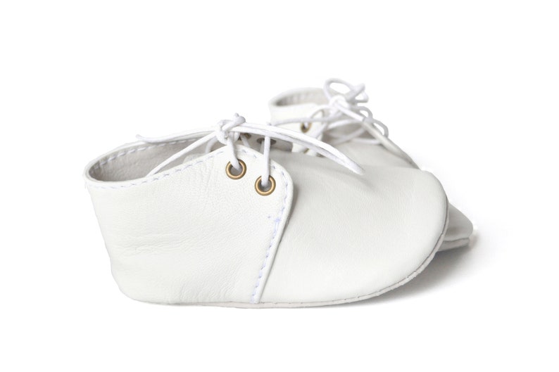Op de afbeelding: Een paar witte leren babyschoenen met veters. De schoenen hebben een klassiek ontwerp met een ronde neus en kleine messing oogjes. De veters zijn wit en geknoopt.