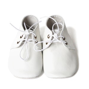 Op de afbeelding: Een paar witte babyschoenen met veters. De schoenen hebben een klassiek ontwerp met een ronde neus en vetergaten. De veters zijn wit en in een strik geknoopt. De schoenen zijn gemaakt van een zacht materiaal.