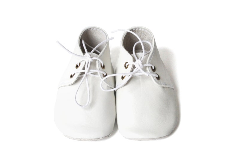 Op de afbeelding: Een paar witte babyschoenen met veters. De schoenen hebben een klassiek ontwerp met een ronde neus en vetergaten. De veters zijn wit en in een strik geknoopt. De schoenen zijn gemaakt van een zacht materiaal.