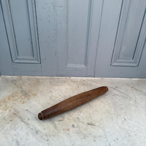 Può includere: Un mattarello in legno con finitura marrone scuro. Il mattarello è cilindrico con estremità arrotondate. È appoggiato su una superficie di marmo davanti a una porta azzurra.