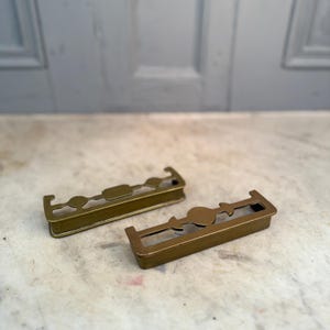 Puede incluir: Dos llaveros de metal color latón con recortes decorativos. Cada soporte rectangular presenta un diseño central y lados abiertos para la inserción de llaves. Los soportes están colocados sobre una superficie clara.