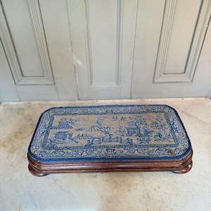 May include: A rectangular footstool with a detailed blue and beige tapestry top depicting an Asian landscape. The footstool has a dark wooden frame and short, curved legs, presenting a classic design. The measurements are in centimeters.
