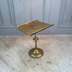 Könnte beinhalten: Ein Vintage-Notenständer aus Messing. Der Ständer hat eine rechteckige, geneigte Oberseite mit einem dekorativen Emblem. Er hat einen runden Sockel und einen verzierten Stiel. Die Oberseite ist horizontal gerillt. Der Ständer ist aus Messing und poliert.