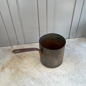 Könnte beinhalten: Ein antiker Kupfertopf mit einem langen, schwarzen Eisenhenkel. Der Topf hat ein abgenutztes, rustikales Aussehen mit einer dunklen Patina.