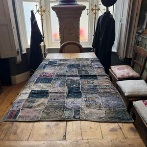 May include: A rectangular patchwork textile runner featuring a variety of square fabric panels in shades of blue, grey, and muted earth tones. The runner displays intricate embroidery, small circular mirrors, and diverse patterns, laid flat on a wooden table.