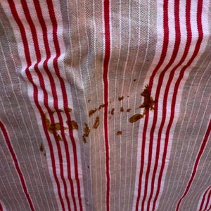 May include: Close-up of a fabric with vertical red and white stripes. The fabric has brown stains in the lower right and center. The fabric appears to be slightly wrinkled.