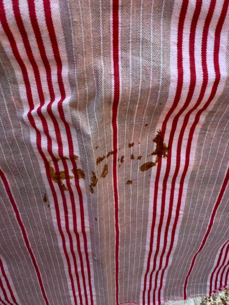 May include: Close-up of a fabric with vertical red and white stripes. The fabric has brown stains in the lower right and center. The fabric appears to be slightly wrinkled.