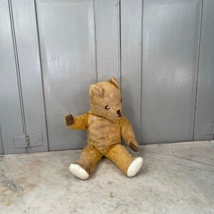 Puede incluir: Un oso de peluche vintage de color beige con ojos marrones y una nariz cosida se sienta sobre una superficie de m&aacute;rmol blanco. El oso tiene patas de fieltro blanco y le falta algo de pelaje.