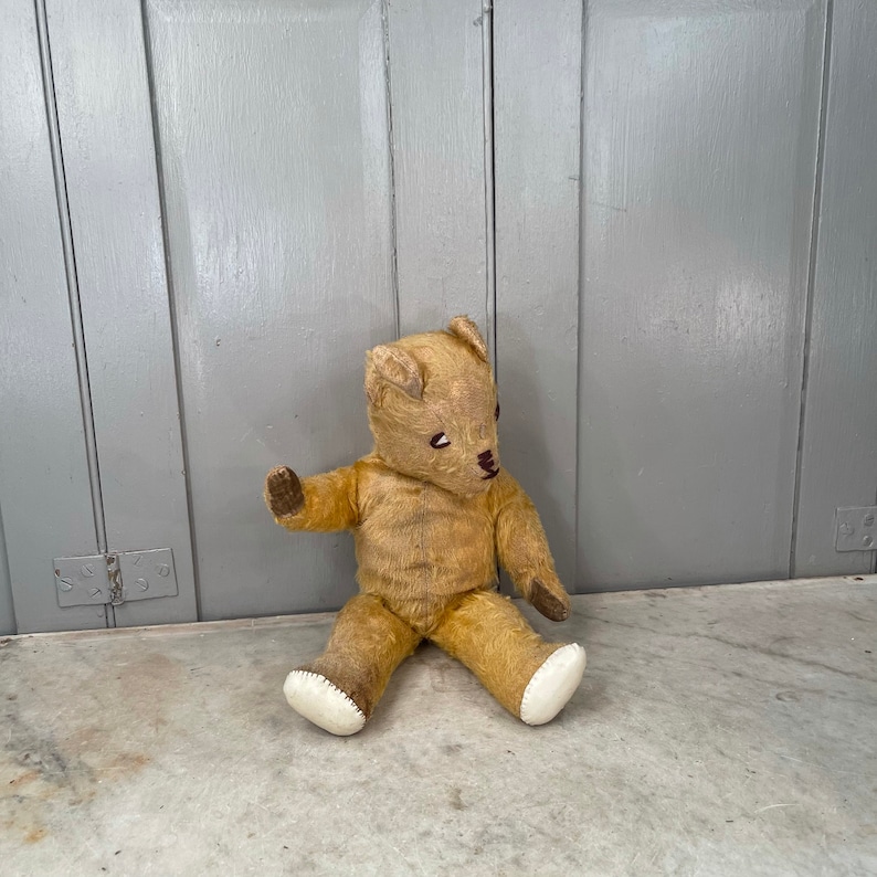 Puede incluir: Un oso de peluche vintage de color beige con ojos marrones y una nariz cosida se sienta sobre una superficie de m&aacute;rmol blanco. El oso tiene patas de fieltro blanco y le falta algo de pelaje.