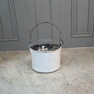 Op de afbeelding: Witte metalen emmer met een zwarte metalen handgreep. De emmer heeft een versleten, verweerde uitstraling en is licht beschadigd.