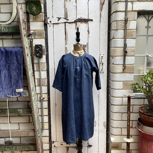 May include: A navy blue linen dress with a stand-up collar and a button closure. The dress is displayed on a black mannequin stand against a white wooden door.