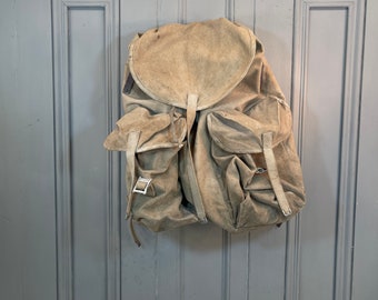 May include: A tan canvas backpack with two large front pockets and a flap closure. The backpack is hanging on a gray door.
